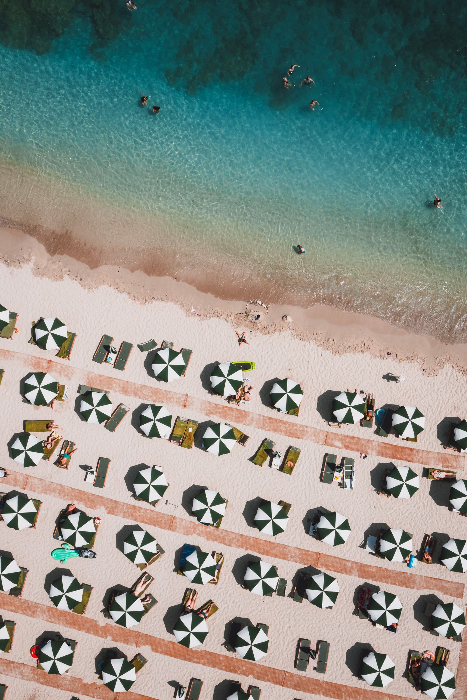 Rows of striped parasols from directly above, the sea cutting a diagonal across the sand.