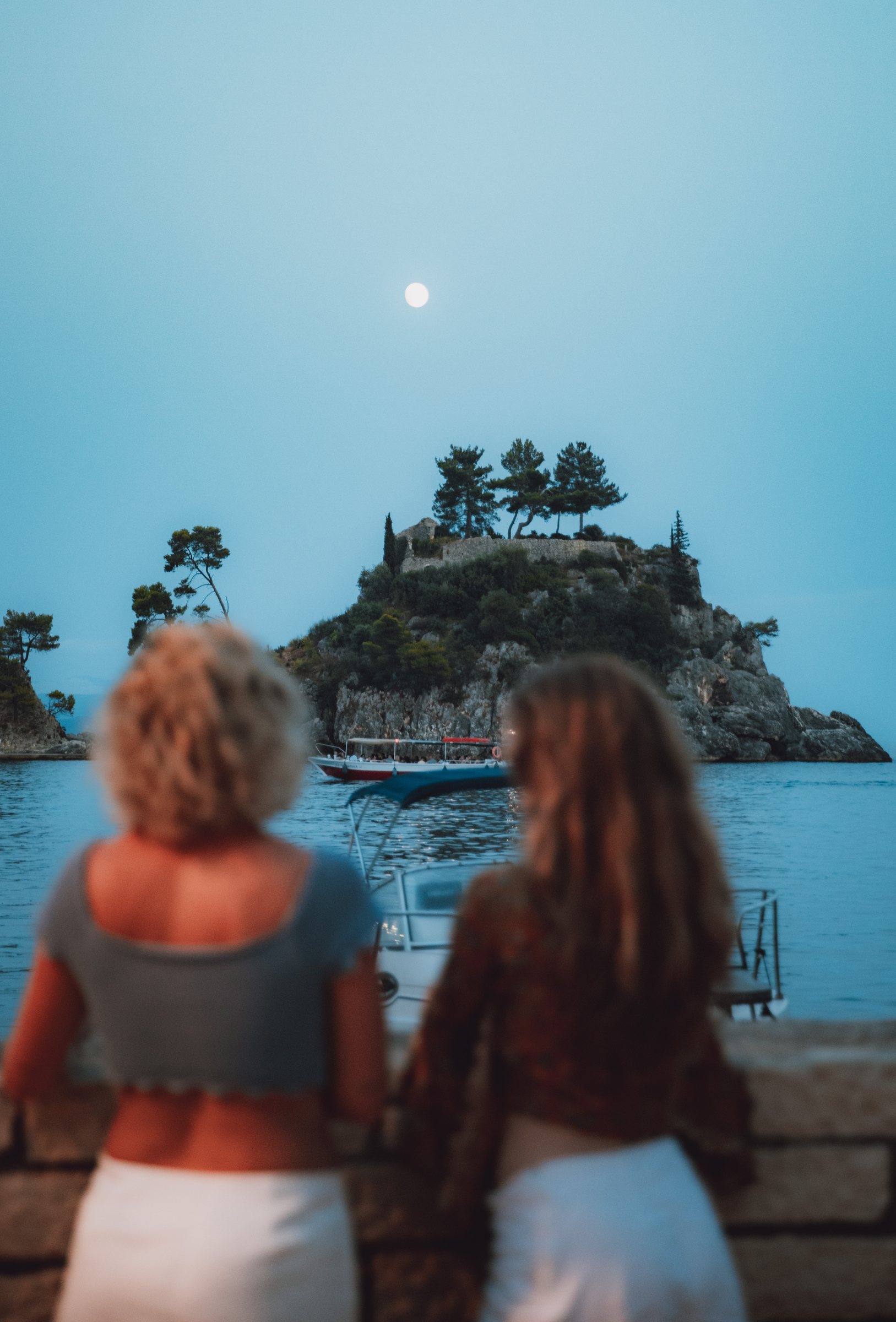 Two figures watching the island at dusk, the moon already up.